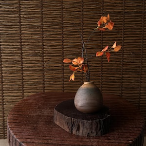 Op de afbeelding: Een kleine, ronde keramische vaas met horizontale ribbels, met takken met oranje bladeren. De vaas staat op een donkere houten voet, geplaatst op een grotere, ronde houten tafel. De achtergrond is een geweven, bruine muur.
