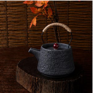 Peut inclure: Une théière en céramique gris foncé texturée, avec une anse tressée et un petit bouton rouge sur le couvercle. La théière est posée sur une surface en bois, avec des feuilles orange en arrière-plan. La théière a un bec verseur.