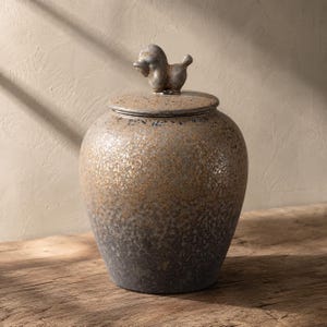 Può includere: Un barattolo in ceramica con coperchio caratterizzato da una piccola figura animale scolpita sulla parte superiore. Il barattolo presenta una finitura testurizzata e maculata che sfuma da un tono marrone dorato chiaro a un grigio scuro.