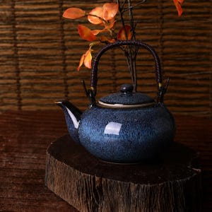 Può includere: Una teiera in ceramica blu con finitura smaltata maculata. Ha un manico e un beccuccio marrone scuro e un coperchio abbinato. La teiera è appoggiata su una superficie di legno, con fiori arancioni sullo sfondo.