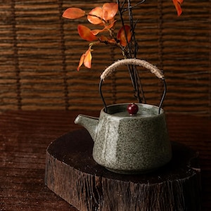 Peut inclure: Petite théière en céramique verte tachetée, avec un bouton rouge foncé sur le couvercle. La théière a une anse tressée et un bec verseur. Elle repose sur une surface en bois, avec des feuilles et des branches orange en arrière-plan.