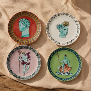 Może przedstawiać: Cztery okrągłe ceramiczne talerze z klasycznymi postaciami mitologicznymi. Czerwony talerz przedstawia zielone popiersie, biały zieloną głowę z muszlą, różowy postać z trójzębem, a zielony talerz ukazuje siedzącą postać.