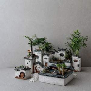 Puede incluir: Colección de casas en miniatura de cerámica blanca con detalles en azul oscuro, cada una con pequeñas plantas y suculentas. Las casas tienen detalles arquitectónicos, creando una escena de pueblo en miniatura.