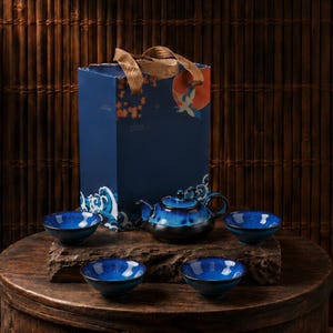 May include: A blue ceramic tea set with a matching teapot and four teacups, presented on a wooden surface. The set has a glossy finish with dark blue and black accents. A navy blue gift box with a crane design is in the background.