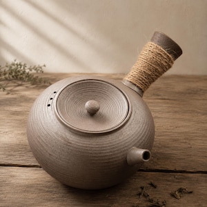 May include: A round, textured, light brown ceramic teapot with a side handle wrapped in natural jute twine. The teapot features a circular lid with a small knob, a short spout, and three small steam holes near the lid opening on the main body.