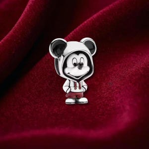 Puede incluir: Un charm de plata con Mickey Mouse con sudadera con capucha y pantalones cortos. El charm tiene detalles en blanco y negro en las orejas y la cara, con pantalones cortos rojos y una sudadera con capucha blanca. El charm est&aacute; sobre un fondo rojo oscuro.