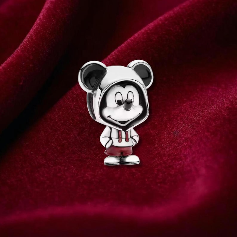 Puede incluir: Un charm de plata con Mickey Mouse con sudadera con capucha y pantalones cortos. El charm tiene detalles en blanco y negro en las orejas y la cara, con pantalones cortos rojos y una sudadera con capucha blanca. El charm est&aacute; sobre un fondo rojo oscuro.