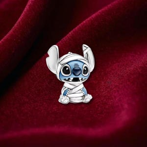 May include: A silver-toned charm featuring a blue and white character in a mummy costume. The charm has large black eyes, a wide grin, and large ears. The character is wrapped in white bandages. The background is a deep red velvet.