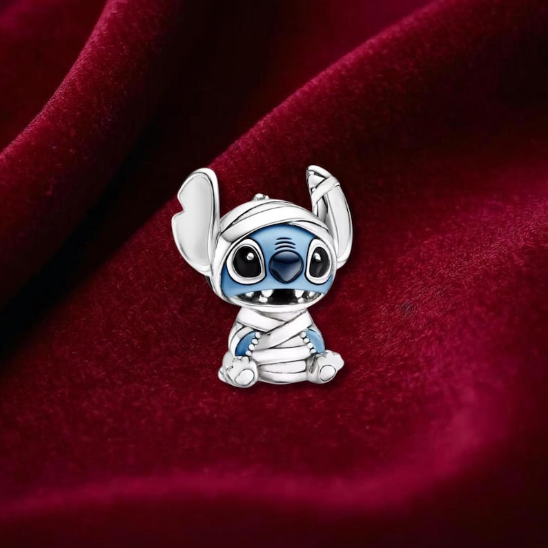May include: A silver-toned charm featuring a blue and white character in a mummy costume. The charm has large black eyes, a wide grin, and large ears. The character is wrapped in white bandages. The background is a deep red velvet.
