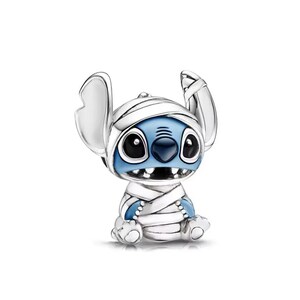 May include: A silver-colored charm featuring a blue alien character wrapped in white bandages, resembling a mummy. The character has large black eyes, a dark blue nose, and a wide grin. The charm is a collectible item.