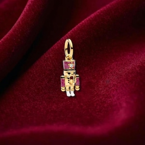 May include: A gold-coloured charm in the shape of a nutcracker soldier. The charm has a red hat and accents, with a star-shaped gem on the hat. The soldier's uniform features red and white details, and white boots. A festive holiday-themed charm.