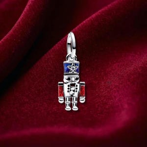 May include: A silver nutcracker charm featuring a blue hat with a central star and a clear stone, red glitter accents on the arms, and black detailing on the face. The charm includes a circular bail at the top for attachment to a bracelet or necklace.