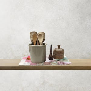 May include: A light gray ceramic crock filled with various wooden spoons, a small dark wooden pestle, and a round wooden lidded container. These items are arranged on a rectangular fabric mat featuring a patchwork of pink and white squares.