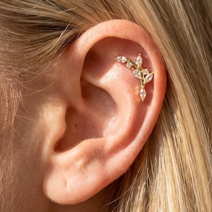 May include: A gold-toned ear cartilage piercing jewelry piece featuring a curved, branch-like design adorned with multiple marquise-cut and round-cut clear stones. The decorative element follows the natural curve of the upper ear cartilage.