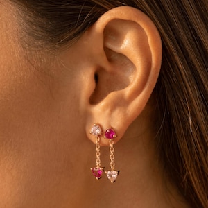 May include: Gold-toned earrings with heart-shaped gemstones. Each earring features a small heart stud and a dangling chain with a matching heart-shaped gemstone. The gemstones are pink and clear.