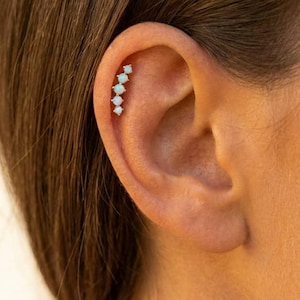 May include: A curved ear climber earring featuring five small, round, iridescent white opal stones set in a vertical row along the upper cartilage of the ear. The stones are arranged in a graduated size pattern, following the natural curve of the ear.
