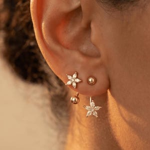 May include: Close-up of three gold earrings. One is a small stud, another is a small ball, and the third is a flower-shaped earring with a dangling design. The earrings are set against a warm, neutral background.