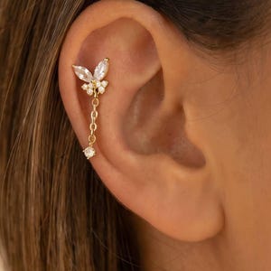 May include: A gold-colored ear piercing featuring a butterfly design with clear, sparkling stones. A delicate chain with a single dangling stone extends from the butterfly. The jewelry is set against the backdrop of a person's ear.
