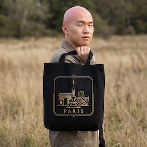Peut inclure: Sac fourre-tout noir avec une illustration dorée des monuments parisiens, dont la Tour Eiffel, l'Arc de Triomphe et le musée du Louvre. Le mot "PARIS" est imprimé en dessous. Le sac a deux anses noires.