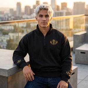 Peut inclure: Sweat-shirt noir à fermeture éclair quart de col avec un écusson doré sur la poitrine et la manche gauche. L'écusson représente une couronne et la lettre "T". Le sweat-shirt a un col montant et une fermeture éclair. Le personne porte un jean bleu.