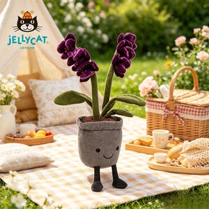 K&ouml;nnte beinhalten: Ein l&auml;chelnder Pl&uuml;sch-Blumentopf mit einer lila Blume und gr&uuml;nen Bl&auml;ttern. Der graue Topf hat schwarze Beine. Der Hintergrund zeigt einen Picknickkorb, Essen und das Jellycat London Logo.