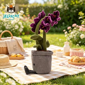 K&ouml;nnte beinhalten: Ein Pl&uuml;sch-Orchidee in einem grauen Topf mit schwarzen Beinen, auf einer Picknickdecke. Die Blume hat gr&uuml;ne Stiele und Bl&auml;tter. Das Jellycat London-Logo ist sichtbar. Im Hintergrund ein Korb, Essen und ein Strohhut.