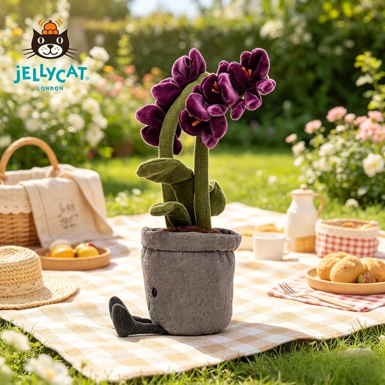 K&ouml;nnte beinhalten: Ein Pl&uuml;sch-Orchidee in einem grauen Topf mit schwarzen Beinen, auf einer Picknickdecke. Die Blume hat gr&uuml;ne Stiele und Bl&auml;tter. Das Jellycat London-Logo ist sichtbar. Im Hintergrund ein Korb, Essen und ein Strohhut.
