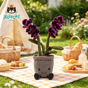 K&ouml;nnte beinhalten: Ein kuscheliger, l&auml;chelnder Blumentopf mit lila Bl&uuml;ten und gr&uuml;nen Stielen. Der Topf ist grau und hat schwarze F&uuml;&szlig;e. Das Jellycat-Logo ist im Hintergrund zu sehen, zusammen mit Picknickk&ouml;rben und Essen auf einer karierten Decke.
