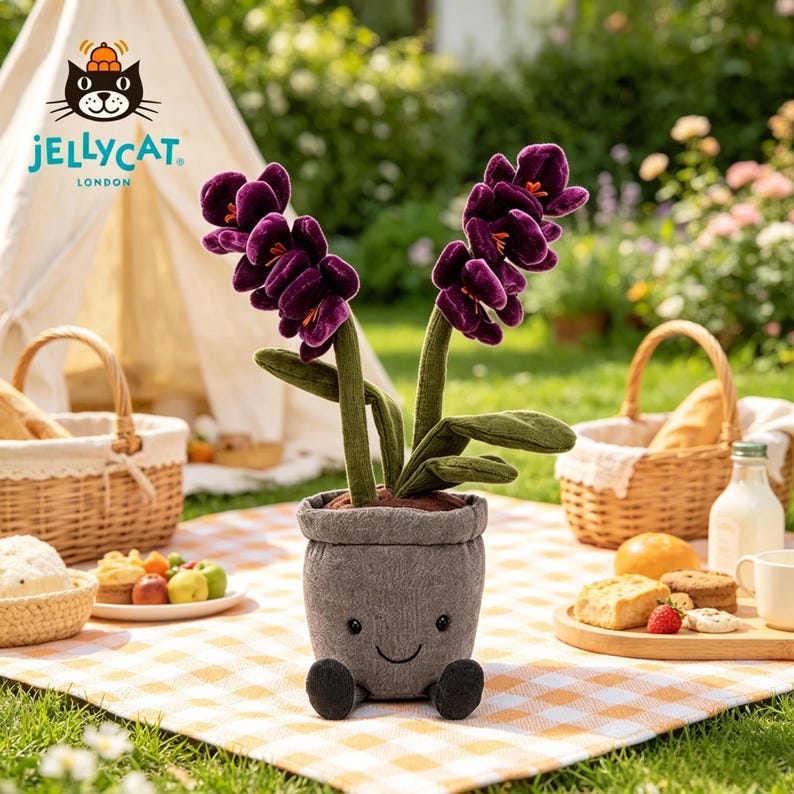 K&ouml;nnte beinhalten: Ein kuscheliger, l&auml;chelnder Blumentopf mit lila Bl&uuml;ten und gr&uuml;nen Stielen. Der Topf ist grau und hat schwarze F&uuml;&szlig;e. Das Jellycat-Logo ist im Hintergrund zu sehen, zusammen mit Picknickk&ouml;rben und Essen auf einer karierten Decke.