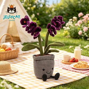 K&ouml;nnte beinhalten: Eine lila Pl&uuml;sch-Orchideenpflanze in einem grauen Topf mit einem l&auml;chelnden Gesicht und kleinen schwarzen F&uuml;&szlig;en. Die Pflanze steht auf einer gelb-wei&szlig; karierten Picknickdecke. Ein Picknickkorb, ein Hut und Essen sind ebenfalls zu sehen.