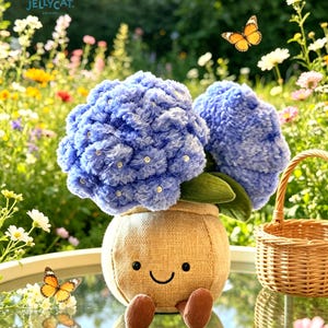 K&ouml;nnte beinhalten: Ein Arrangement aus blauen Pl&uuml;sch-Hortensienbl&uuml;ten in einem l&auml;chelnden Jutesack-Topf mit braunen F&uuml;&szlig;en. Das Arrangement steht auf einem Glastisch mit einem kleinen Weidenkorb und Schmetterlingen im Hintergrund. Das Jellycat-Logo befindet sich oben links.