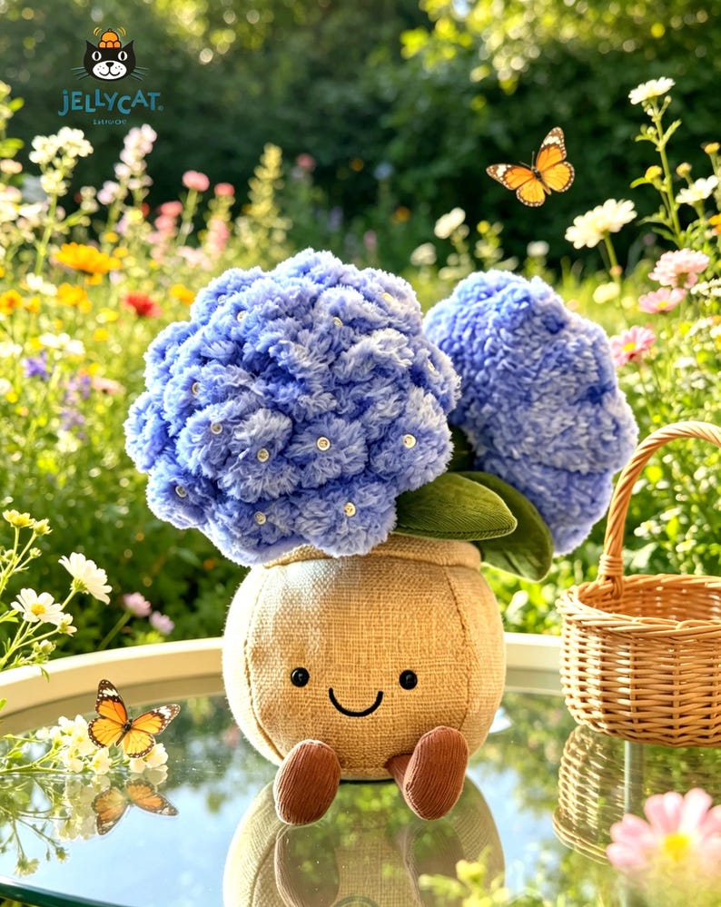 K&ouml;nnte beinhalten: Ein Arrangement aus blauen Pl&uuml;sch-Hortensienbl&uuml;ten in einem l&auml;chelnden Jutesack-Topf mit braunen F&uuml;&szlig;en. Das Arrangement steht auf einem Glastisch mit einem kleinen Weidenkorb und Schmetterlingen im Hintergrund. Das Jellycat-Logo befindet sich oben links.