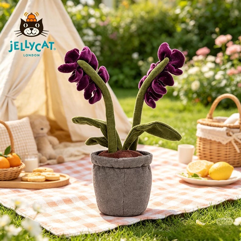 K&ouml;nnte beinhalten: Eine Pl&uuml;sch-Orchideenpflanze in einem grauen Topf. Die Pflanze hat zwei Stiele mit tiefvioletten Bl&uuml;ten und gr&uuml;nen Bl&auml;ttern. Der Topf steht auf einer rot-wei&szlig; karierten Decke. Das Jellycat London Logo ist sichtbar.