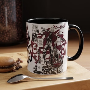 May include: White ceramic mug with a black interior and handle. The mug features a manga-style illustration of a character with the text "Devil May Cry" and panels of text. The mug is on a wooden surface with a spoon and a pastry.