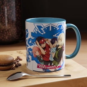 May include: A white ceramic mug with a blue interior and handle, featuring a colorful anime illustration of several characters. The mug has the text "Hata Raku Maou" at the bottom. The mug is on a wooden surface.