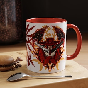 May include: White ceramic mug with a red interior and handle. The mug features a graphic of a character in a red suit and hat holding two guns, with a red and yellow design on the sides. The mug is on a wooden surface.