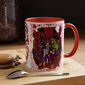May include: A white ceramic mug with a red interior and handle, featuring a graphic illustration of two anime characters sitting on a throne, surrounded by red ornate patterns and Japanese text characters printed on the front surface.