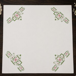 May include: A square white fabric cloth featuring four identical cross-stitch patterns in the corners. Each pattern depicts a central teapot in green and pink thread, flanked by two matching teacups, creating a symmetrical tea-themed design.