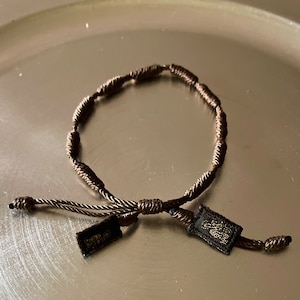 May include: A brown braided bracelet with a knotted closure and two small brown tags. The tags have gold lettering.