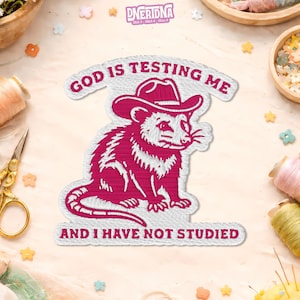 May include: Embroidered patch featuring a pink possum wearing a cowboy hat. The text "GOD IS TESTING ME" arches above the possum, with "AND I HAVE NOT STUDIED" below. The patch is surrounded by white stitching.