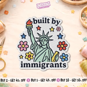 May include: An embroidered patch featuring the Statue of Liberty in light green, holding a torch with an orange flame. The text reads built by immigrants in black. The design includes colorful stars and flowers in pink, yellow, blue, and red.