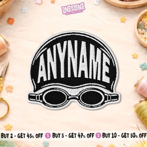 May include: A black and white embroidered patch featuring a swimming cap with the word ANYNAME in bold white capital letters and a pair of swimming goggles underneath. The design is outlined in white thread against a solid black background.
