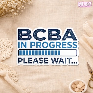 May include: A white sticker with the text "BCBA IN PROGRESS PLEASE WAIT..." in navy blue. The sticker has a progress bar graphic. The sticker is on a beige surface with decorative elements.