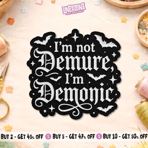 May include: Black embroidered patch with the text "I'm not Demure, I'm Demonic" in white gothic lettering. The patch features bat and star designs. The patch is surrounded by a black border.