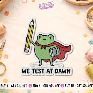 May include: Embroidered patch featuring a green frog wearing a red cape and holding a yellow pencil and a book. The text "WE TEST AT DAWN" is below the frog. The patch has a white border and is likely for clothing or accessories.