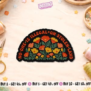 May include: Black patch with the text "NO ONE IS ILLEGAL ON STOLEN LAND" in a semi-circle. The patch features a floral design with orange and yellow flowers. The patch has a black border and is on a light-colored surface.