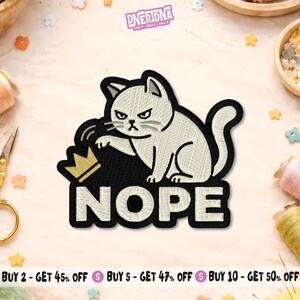 May include: Embroidered patch featuring a grumpy white cat with a black outline, sitting on a black background with the word "NOPE" in white. A gold crown is next to the cat. The patch has the text "DNERIDNA" at the top.