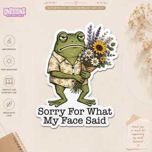 May include: A die-cut sticker featuring a green frog with a grumpy expression wearing a beige floral shirt and holding a bouquet of sunflowers, daisies, and lavender. The text below the frog reads, Sorry For What My Face Said, in black.