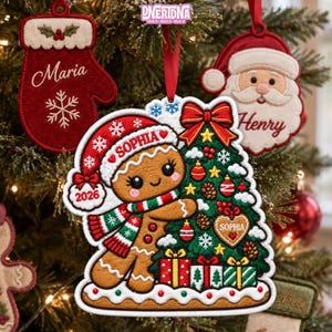 May include: A felt Christmas ornament featuring a gingerbread character wearing a red hat with the name Sophia and the year 2026, hugging a decorated green Christmas tree with gifts, alongside a red mitten ornament and a Santa Claus ornament.
