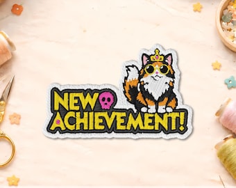 May include: An embroidered patch featuring a calico cat wearing a gold crown and yellow sunglasses. The text NEW ACHIEVEMENT! is stitched in yellow with a black outline, accompanied by a small pink skull icon positioned above the word ACHIEVEMENT.